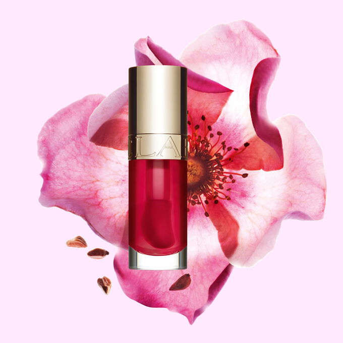 Packshot of Clarins Lip Comfort Oil Fuchsia, placed next to a sweetbriar rose on a light pink background.