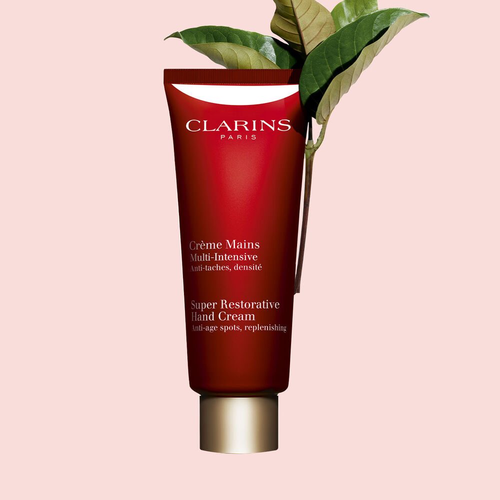 Packshot of a bottle of Clarins Super Restorative Hand Cream close to some harungana leaves on a pink background.